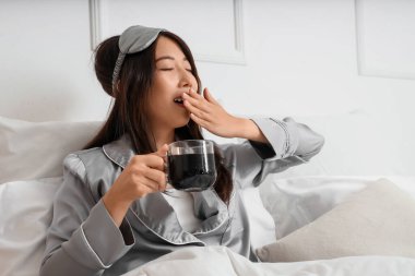 Uyanmış genç Asyalı kadın elinde bir fincan kahveyle yatak odasında esniyor.