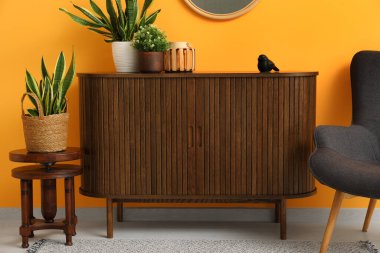 Kahverengi ahşap çekmeceler. Çiçekler, ayna ve sandalye turuncu duvarın yanında.