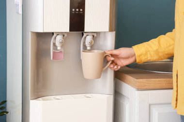 Mavi duvarın yanındaki soğutucudan filtrelenmiş suyla dolduran kadın.