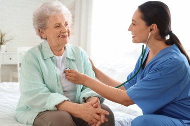 Fizyoterapist yatak odasında steteskoplu yaşlı kadını dinliyor.