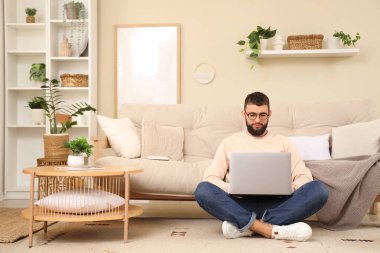 Gözlüklü genç sakallı adam evde dizüstü bilgisayarla çalışıyor.