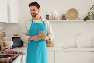 Mutfakta elinde makarna kavanozu ve tavayla yakışıklı sakallı adam.