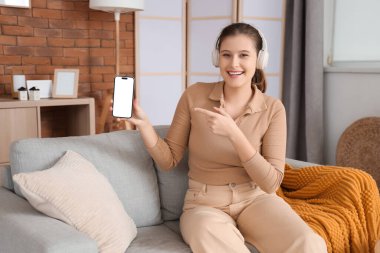 Kulaklıklı genç bir kadın odadaki boş cep telefonunu işaret ediyor.