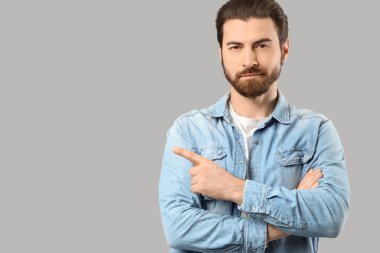 Çapraz kollu sakallı genç adam gri arka plandaki bir şeyi işaret ediyor.
