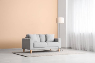 Aydınlık oturma odasının içinde gri kanepe, yer lambası ve penceresi var.