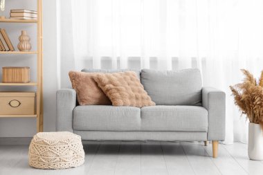 Aydınlık oturma odasının içinde gri kanepe, raf ünitesi ve büyük pencerenin yanında puf, yakın çekim.