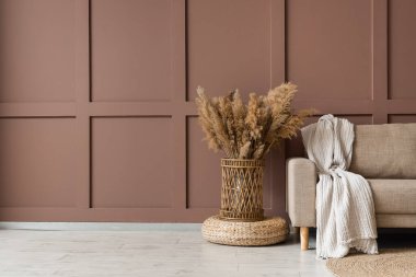 Rahat bir kanepe, hasır puf ve vazo, kahverengi duvarın yanında çimen, oturma odasının içinde, yakın çekim.