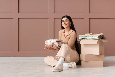 Giysileri ve dolapları olan genç bir kadın kahverengi duvarın yanında oturuyor.