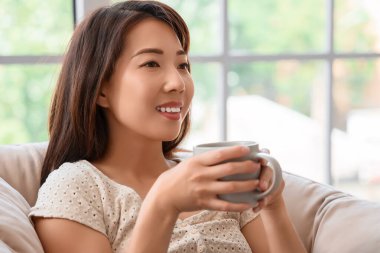 Evdeki koltukta bir fincan kahveli genç Asyalı kadın, yakın plan.