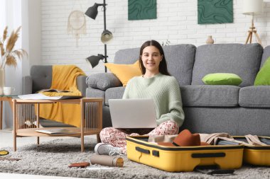 Evdeki bavulun yanında dizüstü bilgisayar kullanan genç bir kadın. Seyahat kavramı