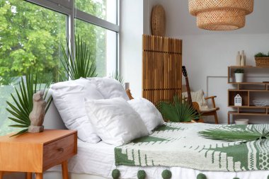 Palmiye dallarıyla döşenmiş rahat bir yatak, komodinli ve yatak odasındaki geniş pencerenin yanında bank.