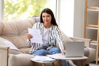 Genç ve güzel iş kadını elinde belgelerle kanepede oturuyor ve ofisteki bilgisayarını kullanıyor.