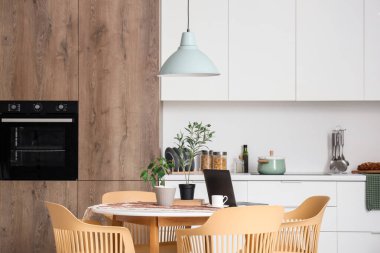 Yemek odasındaki masada bir fincan kahve, ev bitkileri ve modern laptop var.