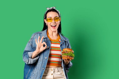 Lezzetli burgeri olan güzel genç bir kadın yeşil arka planda iyi bir jest gösteriyor.
