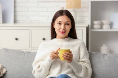 Oturma odasındaki kanepede bir fincan sıcak çayla oturan güzel, genç Asyalı kadın.