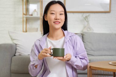 Genç Asyalı kadın oturma odasında oturmuş kahve içiyor.
