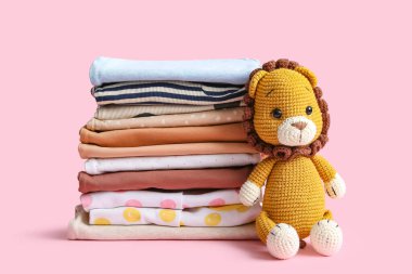 Pembe arka planda oyuncak aslan olan bebek kıyafetleri yığını.