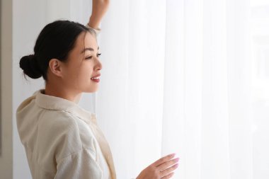 Evdeki ışık perdelerinin yanındaki genç Asyalı kadın.