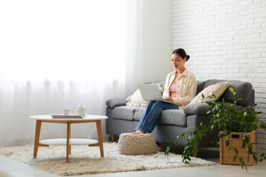 Genç Asyalı kadın evdeki ışık perdelerinin yanındaki kanepede dizüstü bilgisayar kullanıyor.