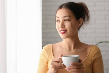 Evinin penceresinin yanında bir fincan kahveli genç Asyalı kadın, yakın plan.