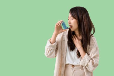 Astım krizi geçiren genç bir Asyalı kadın yeşil arka planda astım spreyi kullanıyor.