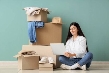 Laptop ve kıyafet kutularıyla yeşil duvarın yanında oturan genç bir kadın.