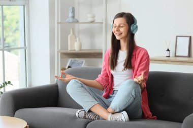 Kulaklıklı genç bir kadın evdeki kanepede meditasyon yapıyor.