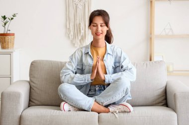 Evdeki kanepede meditasyon yapan genç bir kadın.