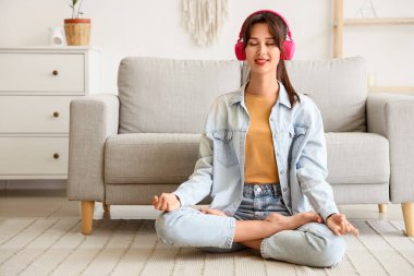 Kulaklıklı genç bir kadın evde halıda meditasyon yapıyor.