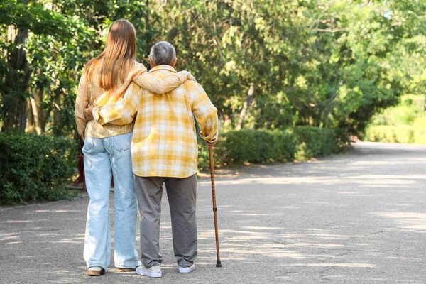 Senior woman with stick and caregiver hugging in park, back view