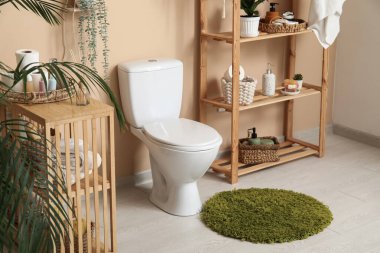 Tuvaletin içinde beyaz klozet, ev bitkisi ve bej duvarın yanında raf ünitesi var.