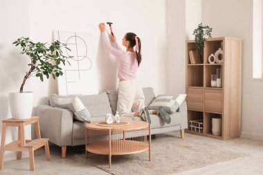 Genç ve güzel bir kadın şık oturma odasındaki beyaz duvara resim asıyor.