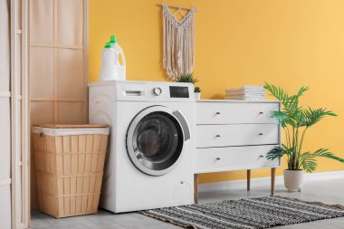 Çamaşır makinesi, çekmece sandığı ve sarı duvarın yanında çamaşır sepeti.