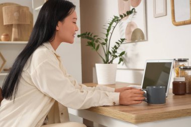 Genç Asyalı kadın kupadan kahve içiyor ve mutfakta dizüstü bilgisayar kullanıyor.