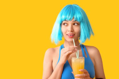 Mayo giymiş genç bir kadın elinde bir bardak portakal suyu ve sarı arka planda pipetler tutuyor.