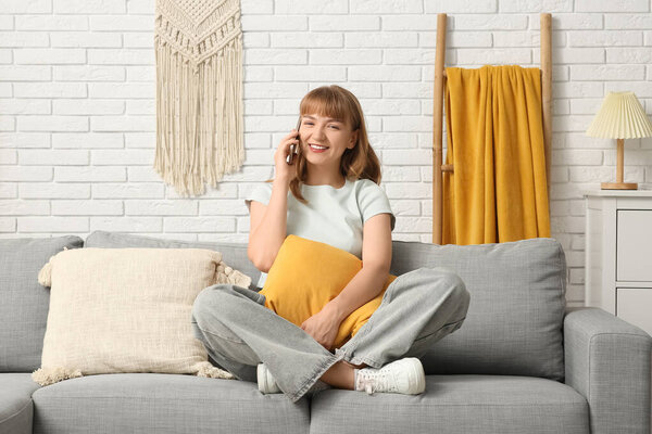 Красивая молодая женщина с подушкой разговаривает по мобильному телефону на сером диване дома