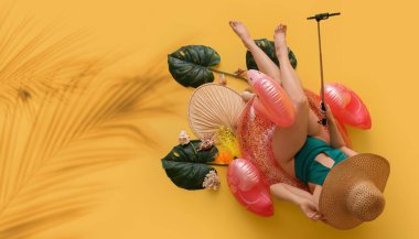 Mayo giymiş genç bir kadın ve şişme yüzükle sarı arka planda tropikal yapraklarla selfie çekiyor.