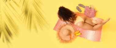 Güneş kremi ve plaj aksesuarları ile sarı arka planda uzanan tropikal yapraklı bir kadın.