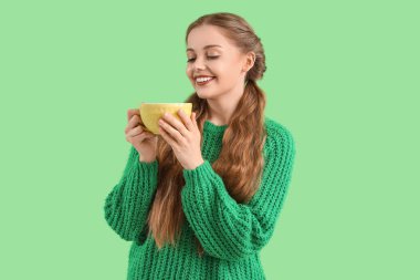 Yeşil arka planda bir fincan sıcak çayla sıcak süveter giyen güzel, mutlu genç bir kadın.