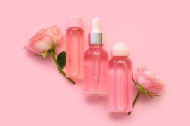 Gül özlü kozmetik yağı şişeleri ve pembe arka planda çiçekler.