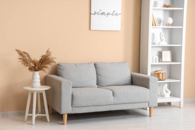 Oturma odasının içindeki bej duvarın yanında gri kanepe ve raf ünitesi.