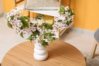 Oturma odasındaki turuncu duvarın yanındaki sehpanın üzerinde çiçek açan dalları olan vazo.