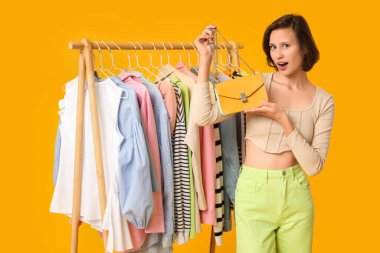 Farklı kıyafetlerle sarı arka planda çanta seçen şaşırmış genç bir kadın.