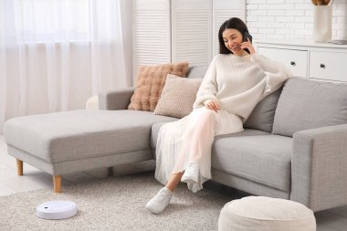 Evde elektrikli süpürgeyle cep telefonuyla konuşan genç bir kadın.