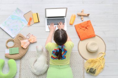 Dizüstü bilgisayarı olan genç bir kadın, sevimli beyaz bir kedi ve evdeki ahşap zeminde farklı seyahat şeyleri.