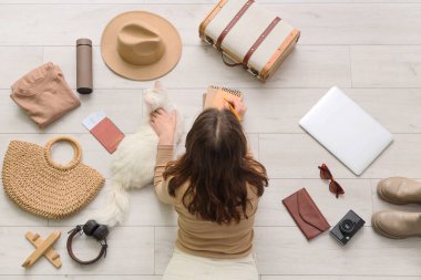 Defterleri olan genç bir kadın, sevimli beyaz bir kedi ve evdeki ahşap zeminde farklı seyahat şeyleri olan bir kadın.