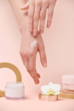 Alçı podyumlu kadın elleri, krema kavanozları ve bej arka planda orkide çiçekleri.