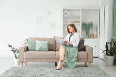 Evdeki gri koltukta dizüstü bilgisayar kullanan güzel genç bir kadın.