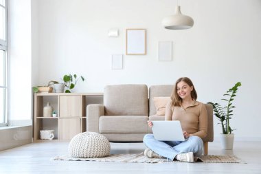 Oturma odasındaki gri kanepenin yanındaki halıda dizüstü bilgisayar kullanan genç bir kadın.