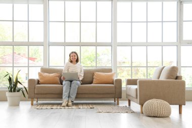 Dizüstü bilgisayarı olan genç bir kadın evdeki pencerenin yanındaki gri koltukta oturuyor.